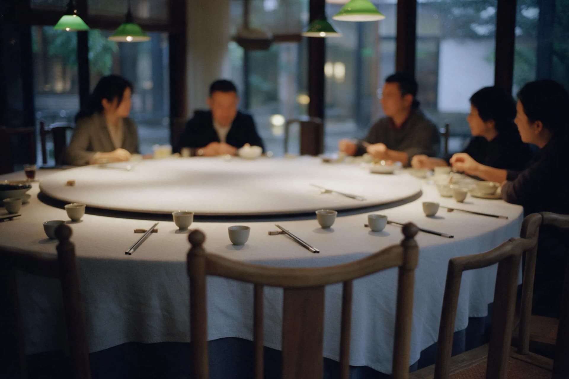 A 35mm photo of an empty guest chair at a round Chinese banquet table with teacups and hosts nearby on a rainy evening