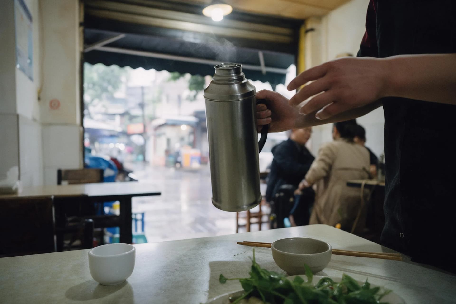 A 35mm travel photo of a Chinese restaurant server pouring hot water from a thermos beside a rinse bowl and cup